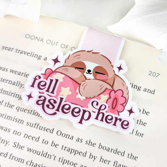 Magnetic bookmark featuring a sleeping sloth cuddling a pink pillow with the phrase “fell asleep here,” clipped to the top of an open book.