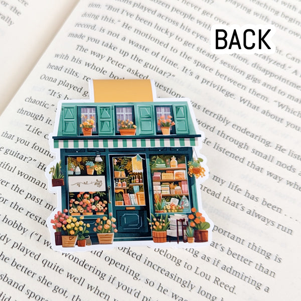 Back view of a magnetic bookmark featuring a colorful bookstore storefront with potted plants, books, and green awning, resting on an open book.