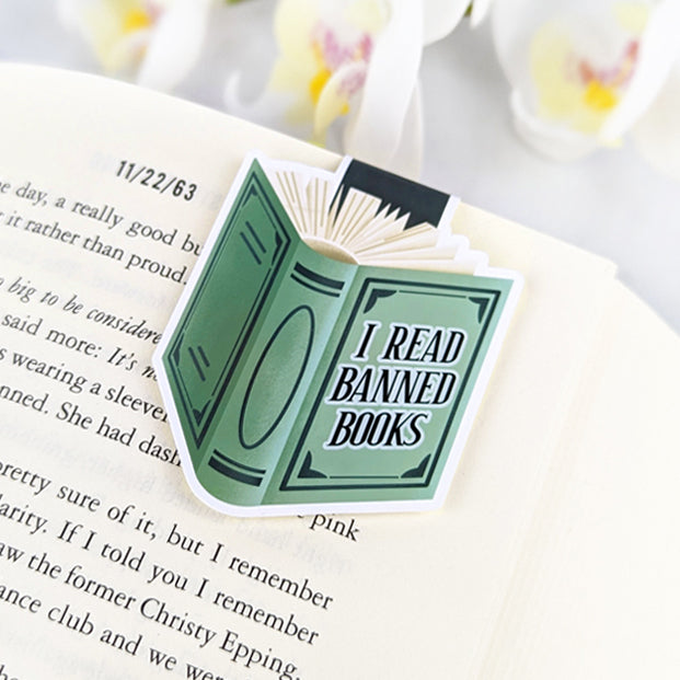 Magnetic bookmark shaped like a green book with the phrase 'I Read Banned Books,' clipped to an open book.