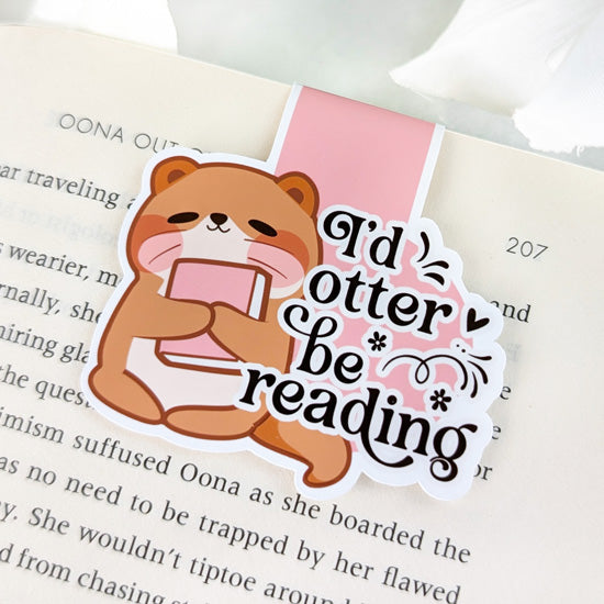 Magnetic bookmark featuring a cute otter hugging a book with the phrase “I’d otter be reading,” clipped to the top of an open book.