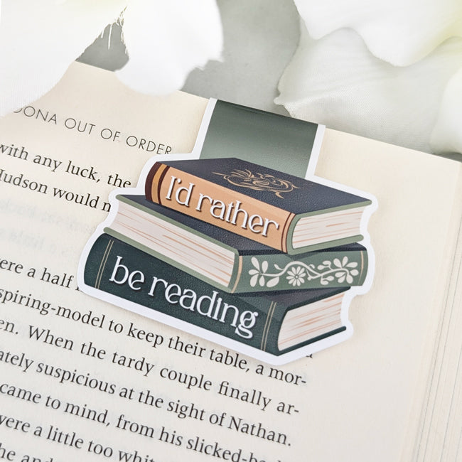 Magnetic bookmark featuring a vintage-style book stack with the phrase 'I'd rather be reading,' clipped to an open book.