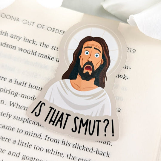 Funny magnetic bookmark with shocked cartoon Jesus and 'Is That Smut?!' text, placed on an open book with white flower decor.