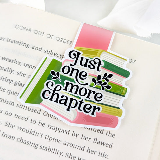 Magnetic bookmark featuring a colorful stack of books with the phrase “Just one more chapter” in bold text, clipped to the top of an open book.
