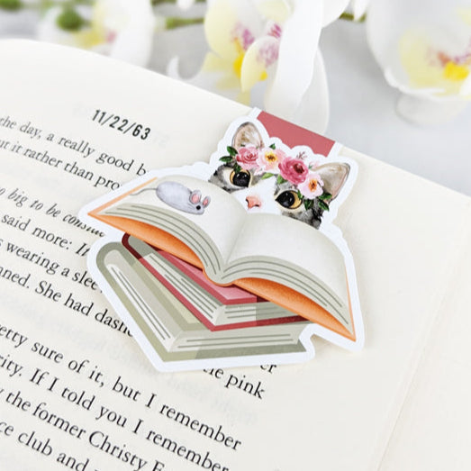 Magnetic bookmark featuring a cat with a flower crown peeking over an open book with a toy mouse, clipped to an open book.