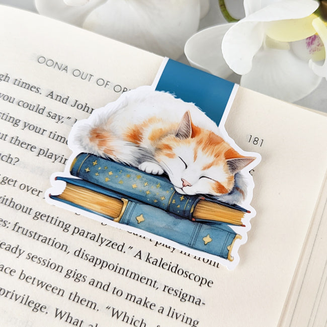 Magnetic bookmark featuring an orange and white cat sleeping on a stack of blue books, clipped to an open book with white flowers nearby.