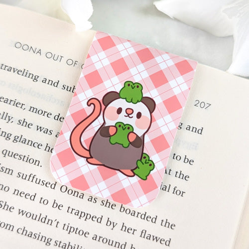 Magnetic bookmark featuring a cute opossum holding and surrounded by green frogs, set against a pink argyle background, clipped to the top of an open book.