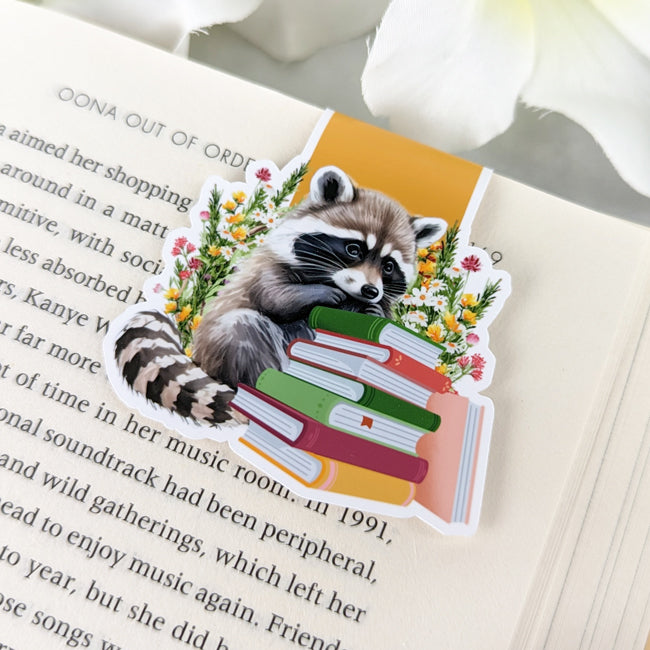 Magnetic bookmark featuring a raccoon with a stack of colorful books and wildflowers, clipped to an open book with white flowers nearby.