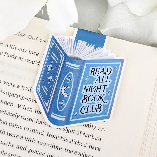 Magnetic bookmark shaped like a blue book with moon and star details and the phrase 'Read All Night Book Club,' clipped to an open book.