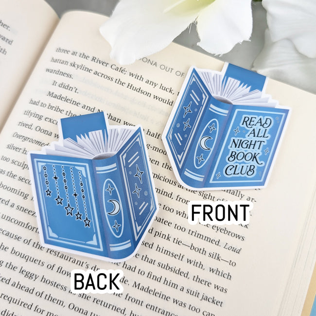 Front and back view of a blue book-shaped magnetic bookmark with moon and star designs and the phrase 'Read All Night Book Club,' clipped to an open book.