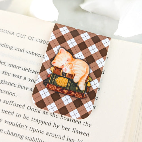 Magnetic bookmark featuring an orange tabby kitten sleeping on a stack of vintage books, set against a brown argyle background, clipped to the top of an open book.