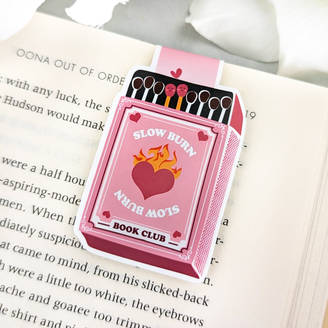 Magnetic bookmark shaped like a pink matchbox with illustrated matches and the phrase "Slow Burn Book Club," clipped to an open book.