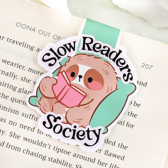 Magnetic bookmark featuring a relaxed sloth reading a pink book on a green cushion with the phrase “Slow Readers Society,” clipped to the top of an open book.
