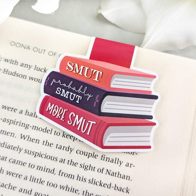 Magnetic bookmark shaped like a stack of colorful books labeled 'Smut,' 'Probably Smut,' and 'More Smut,' clipped to an open book.
