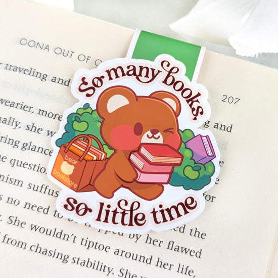 Magnetic bookmark featuring a bear carrying a stack of books with a bag labeled “bear bookstore,” surrounded by greenery, and the phrase “So many books, so little time,” clipped to the top of an open book.