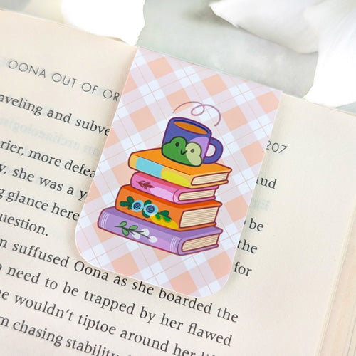 Magnetic bookmark featuring a stack of colorful books topped with a purple mug with a cute green frog design, set against a peach argyle background, clipped to the top of an open book.