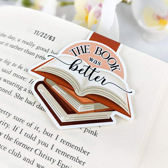 Magnetic bookmark shaped like a stack of books with an open book on top and the phrase 'The book was better,' clipped to an open book.