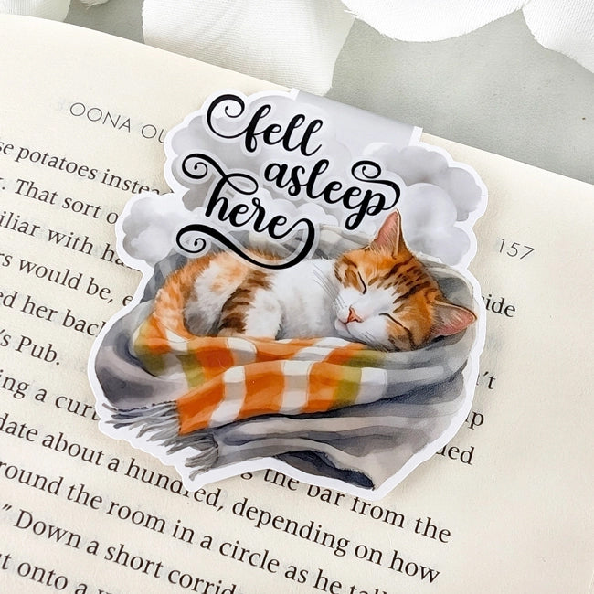 Magnetic bookmark featuring an orange and white cat sleeping in a blanket with 'Fell asleep here' text, clipped to an open book.