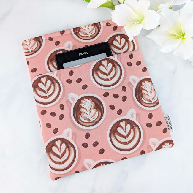 Padded book sleeve, fabric featuring cappuccino and coffee bean illustrations on pink background, white flowers in the background.