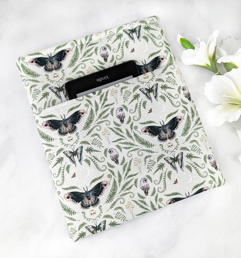 large padded book sleeve with pocket, fabric pattern featuring ferns, moths, mushrooms, owls and skulls on a light green background.