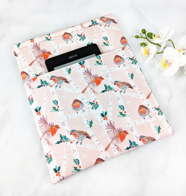 Light pink fabric book sleeve with a birch tree forest pattern featuring robins, holly leaves, and snowflakes, shown holding a hardcover book on a white marble background.