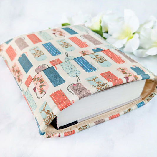 Fabric book sleeve with a whimsical print of illustrated bookmarks in coral, blue, mint, and brown tones on a cream.