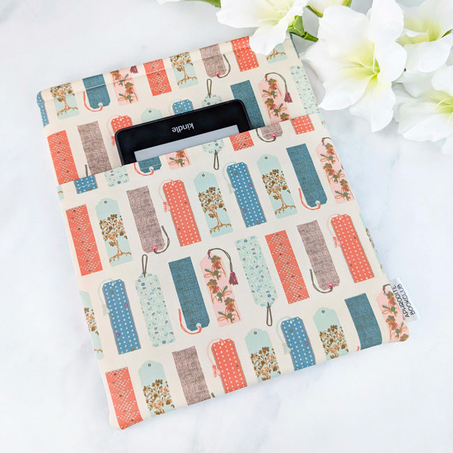 Fabric book sleeve with a whimsical print of illustrated bookmarks in coral, blue, mint, and brown tones on a cream background, shown with a Kindle.