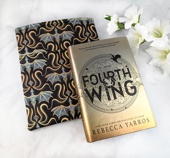 Fabric book sleeve with a bold pattern of gold and gray dragons on a black background, displayed with white flowers on a white surface.