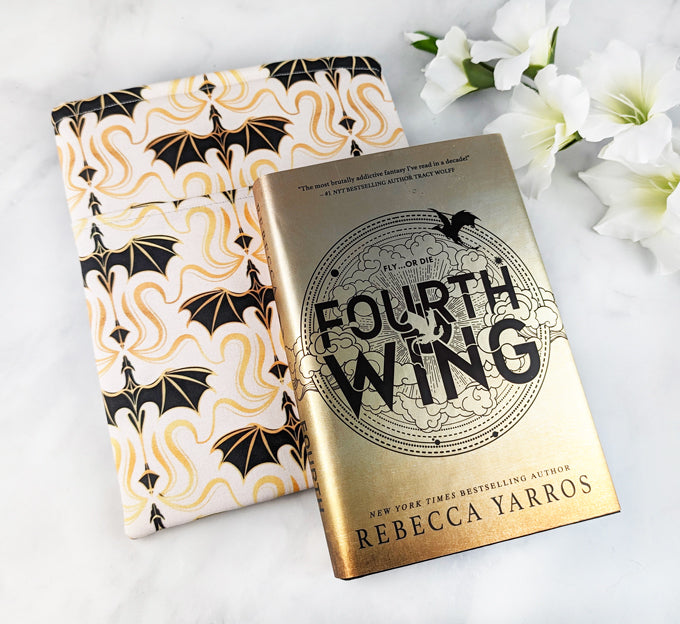 padded large book sleeve with pocket, fabric showcases simple illustrations of flying dragons in a damask style in shades of black, gold and white. next to it there's a book Fourth Wing by Rebecca Yarros.