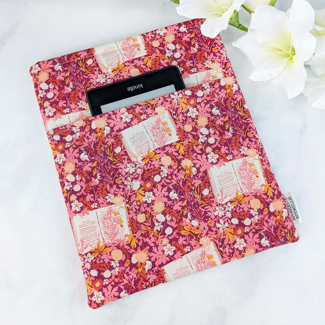 Fabric book sleeve with a cozy vintage-style floral and open book print in soft rose and marigold hues on a berry pink background, styled beside white flowers.