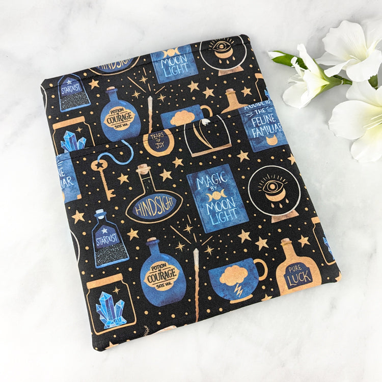large padded book sleeve with pocket, fabric pattern features various witchy items like potions, spell books, crystal balls and more. Colors are blue, yellow gold and white on a black background.