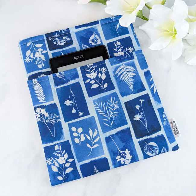 Fabric book sleeve featuring a blue botanical cyanotype-style print with delicate white silhouettes of ferns, leaves, and wildflowers, holding a Kindle, styled with white flowers in the background.