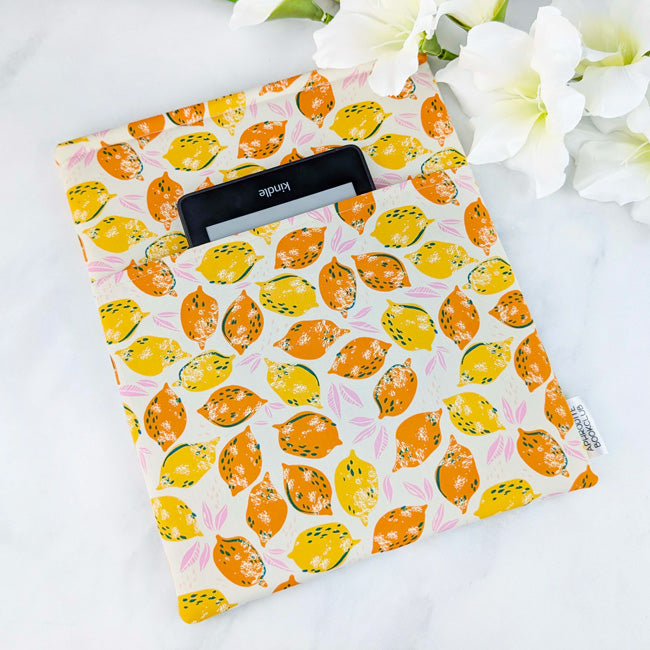 Fabric book sleeve with a vibrant citrus fruit print featuring orange and yellow lemons accented with soft pink leaves on a cream background.