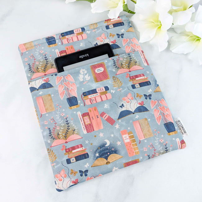 Fabric book sleeve with a whimsical print of stacked books, open pages, plants, butterflies, and stars on a soft gray background, with a Kindle tucked inside.