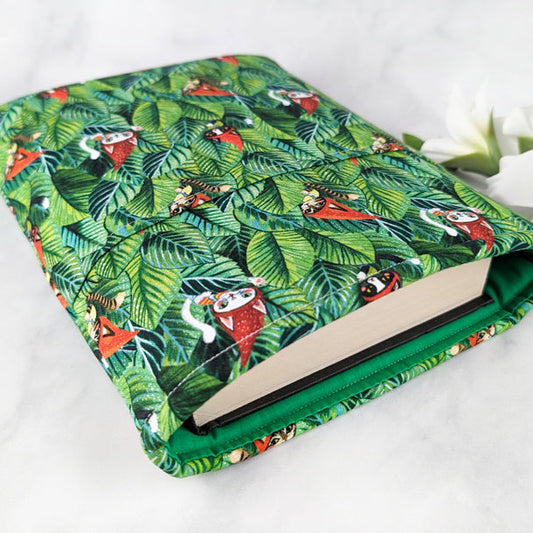 Fabric book sleeve with a jungle leaf pattern featuring cats in red gnome hats, displayed on a white surface with white flowers.