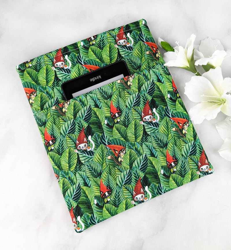 Fabric book sleeve with a jungle leaf pattern featuring cats in red gnome hats, displayed on a white surface with white flowers.