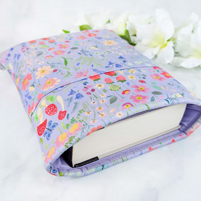 Fabric book sleeve with a light purple background featuring strawberries, mushrooms, and colorful wildflowers, displayed with white flowers on a white surface.