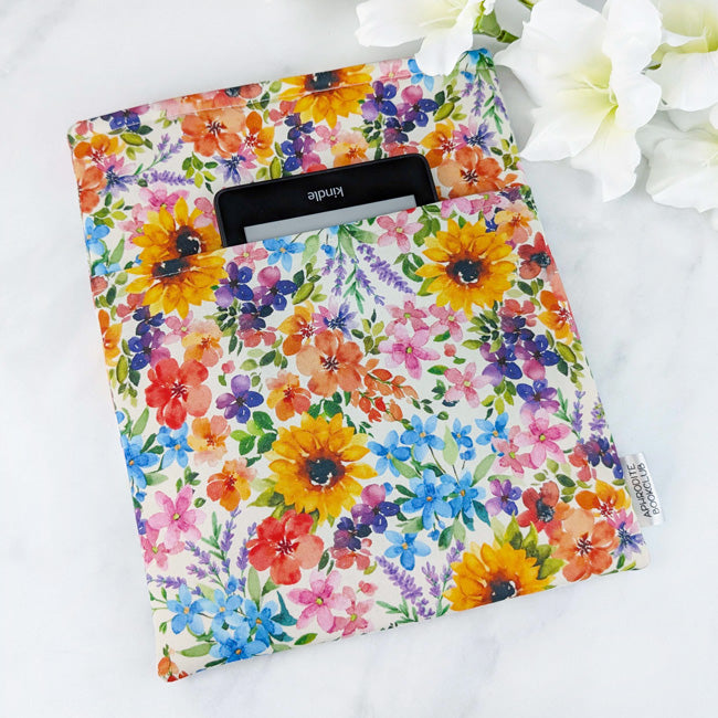 Padded book sleeve, fabric features vibrant floral pattern featuring sunflowers and wildflowers, sleeve is set against a white background with white flowers.