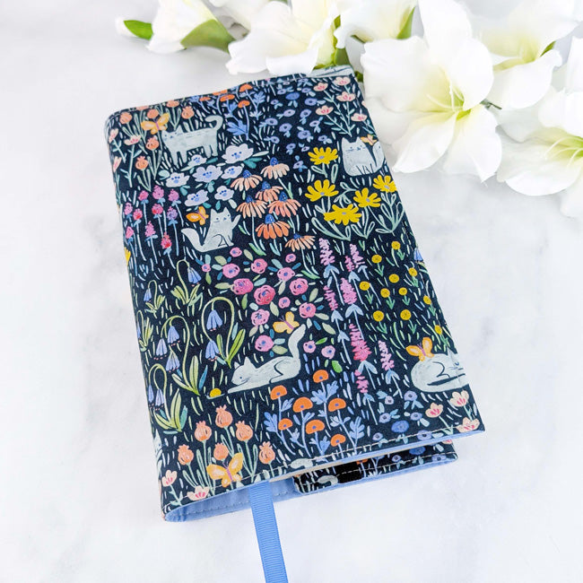 Fabric book cover featuring playful gray cats lounging and hiding among colorful wildflowers on a deep navy background.