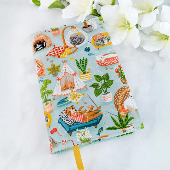 Fabric book cover featuring cozy illustrated cats lounging in hammocks, beds, and tents surrounded by houseplants, against a soft blue background.