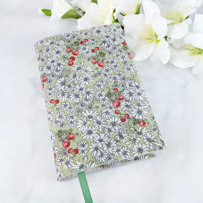 Fabric book cover with a soft green background featuring white daisies and red strawberries, fitted on a book with a green ribbon bookmark, displayed beside white flowers.