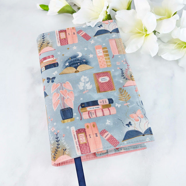 Fabric book cover with a dreamy, whimsical print on soft blue cotton, featuring stacks of pastel books, potted plants, fluttering butterflies, and celestial touches like stars and moons.