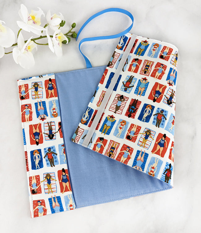 adjustable fabric book cover, fabric pattern features multiple illustrations of women of various races laying on colorful beach towels.