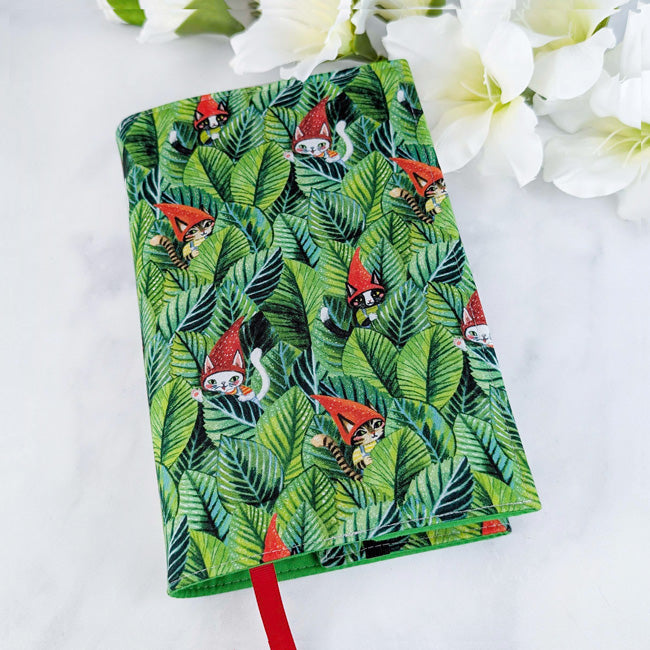 Fabric book cover featuring a pattern of cats wearing red gnome hats hidden among green jungle leaves, fitted on a book with a red ribbon bookmark, displayed beside white flowers.