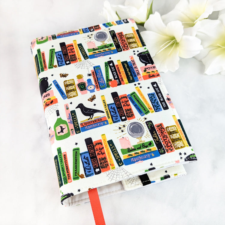 Off-white book cover decorated with colorful illustrated books, potion bottles, ravens, and spell labels in a playful witchy library design.