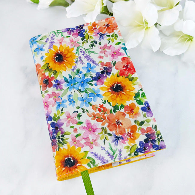 Fabric book cover featuring a vibrant floral pattern with sunflowers, lavender, and assorted wildflowers in bold, cheerful colors, shown with a green ribbon bookmark and white flowers in the background.