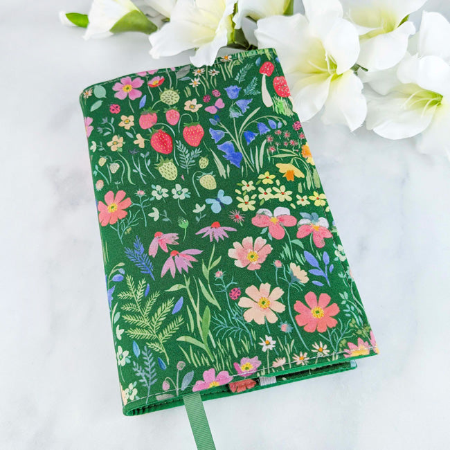 Fabric book cover with a lush meadow-inspired design featuring wild strawberries, colorful flowers, and greenery on a rich forest green background, finished with a matching green ribbon bookmark.