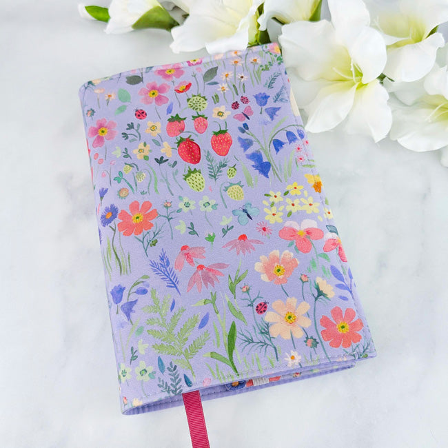 Fabric book cover with a purple background featuring strawberries, flowers, and greenery, fitted on a book with a red ribbon bookmark, displayed beside white flowers.