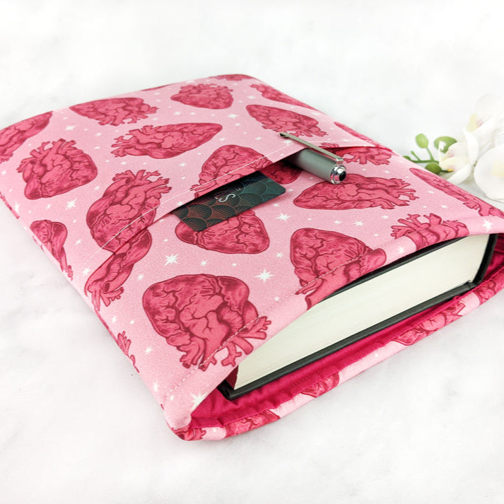 padded book sleeve, fabric shows anatomical red hearts on pink background with tiny white stars between them. In the side pocket there is a pen and a library card.