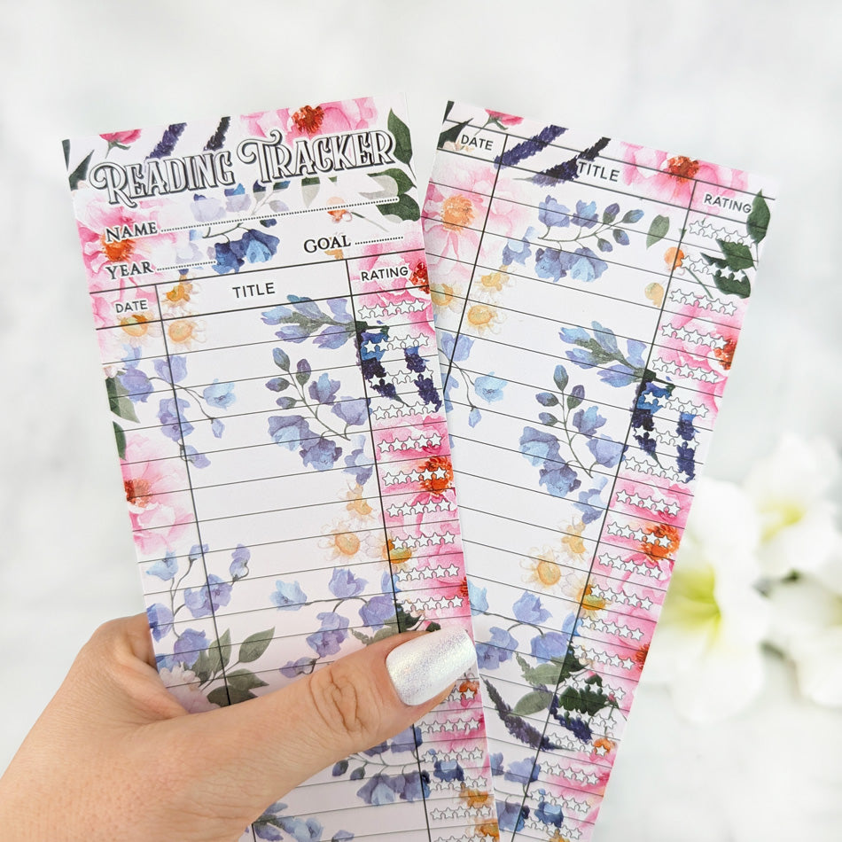 Two floral reading tracker bookmarks with vibrant bouquet designs, held in a hand with silver nail polish.