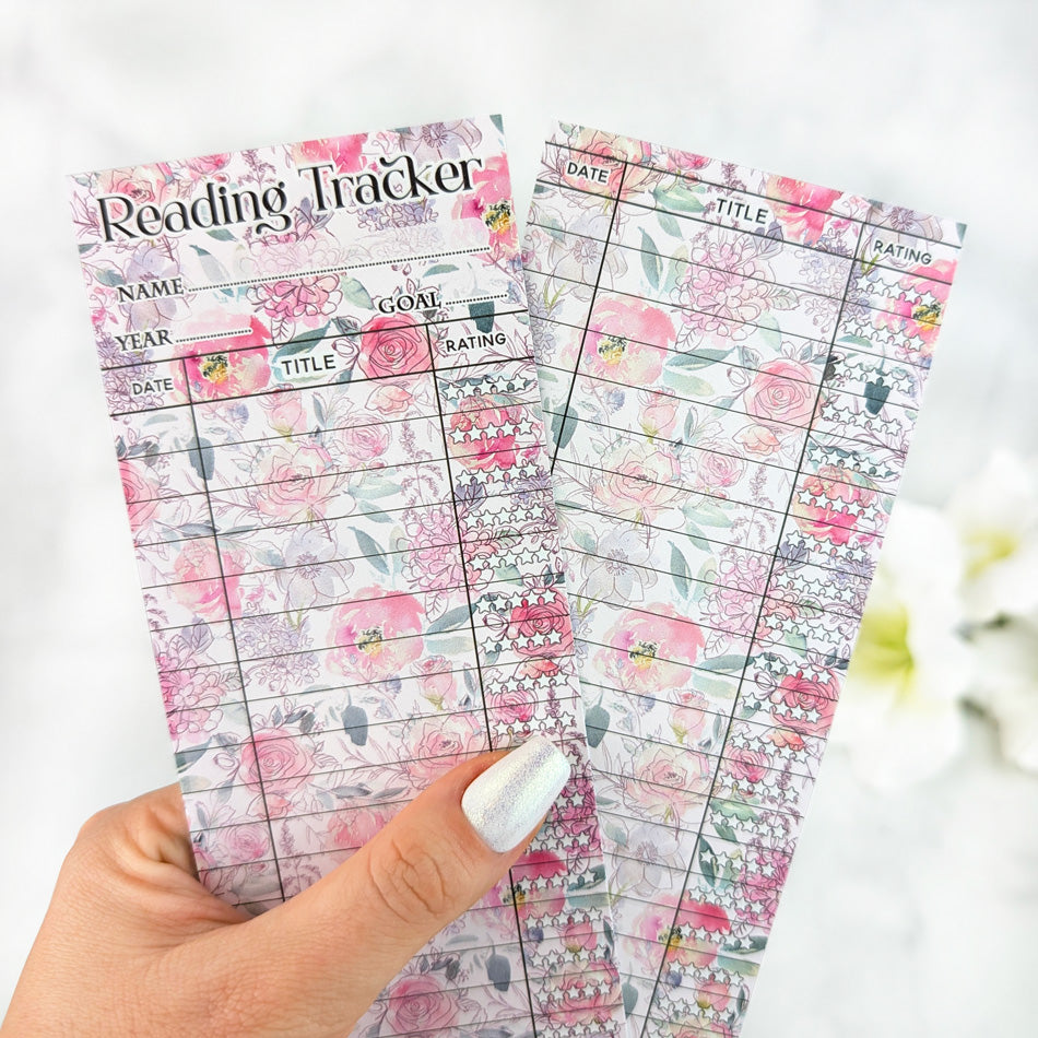 Two pastel floral reading tracker bookmarks with pink rose designs, held in a hand with silver nail polish.